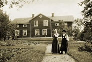 Two women in period-costume ca. 1900 in a field in front of a house.